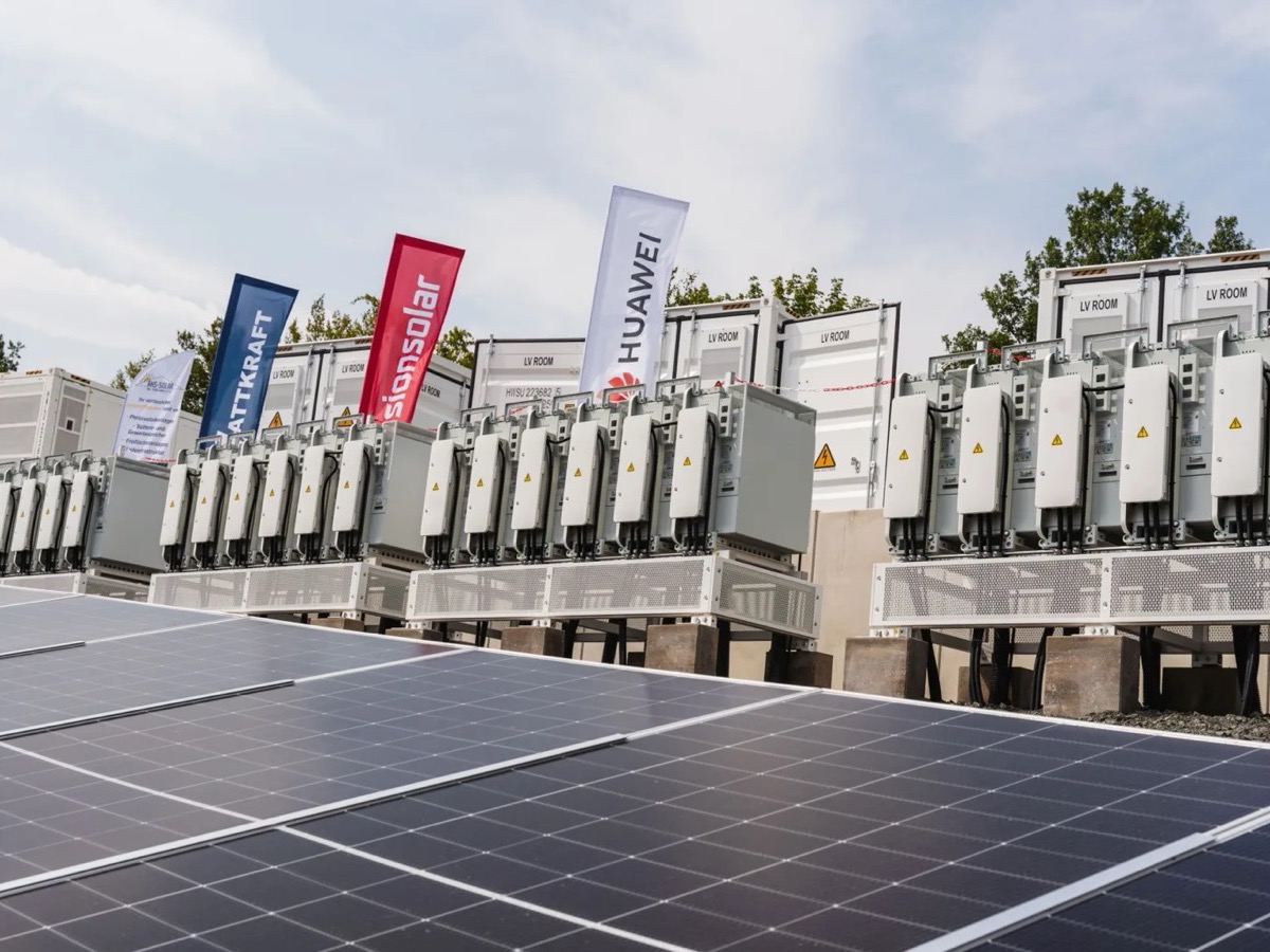 Reihen von Umrichtern vor den Speichercontainern, im Vordergrund Photovoltaikmodule, dazwischen die Fahnen der Partner